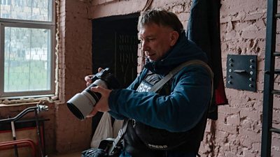 A man in a flak jacket with Press across the front looks at a camera