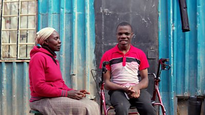 A woman sits beside a young man in a wheelchair - he is smiling. They are in front of a blue corrugated metal wall
