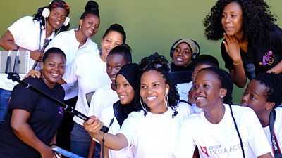 A group of women cluster around a radio microphone. One wears a BBC Media Action T-shirt and they are smiling