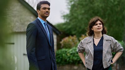 A man in a suit and woman in a smart jacket and blouse stand outdoors, surveying a scene.