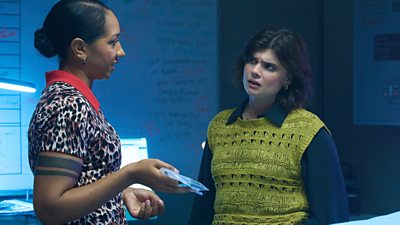 Two women stand discussing something in a room set up for an investigation. One holds what appears to be some bagged evidence.