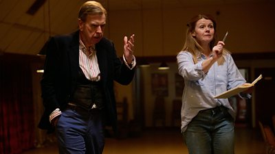 A man and woman stand side by side in a room offering direction to someone off screen