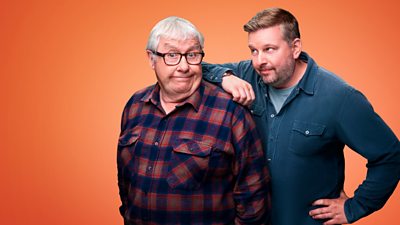 Two men stood in front of an orange background. An older gentleman wears a plaid shirt and glasses. A younger man wears a blue shirt and leans on the older man’s shoulder.
