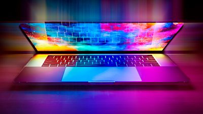 A laptop on a desk in a darkened room. The laptop lid is partially open and colour is shining out from the screen reflecting on the keyboard and giving a colouful aura around the device.