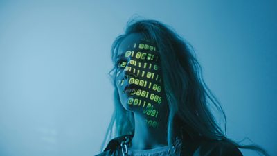 A stark head and shoulders image of a woman on a pale background - computer code is projected onto her face.