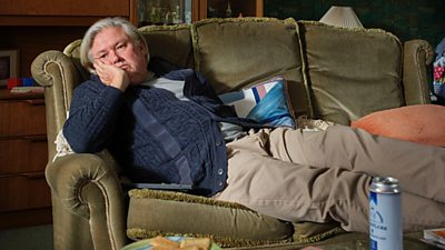 A man slumped in an old fashioned olive green sofa. He is wearing a cardigan and has his head in his hand. There is a beer can on the table in front of him