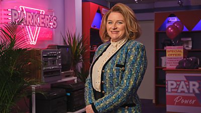 A woman smiling, stood in front of a pink neon sign that reads Parker's. There are also some balloons, a large pot plant and a retro hi-fi in shot
