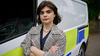 A woman stands by a police van with her arms crossed