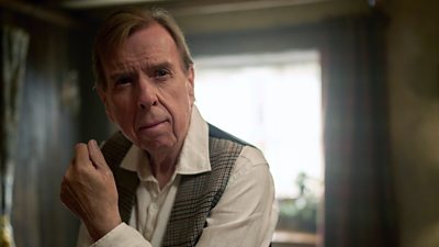 A man in a waistcoat and white shirt looks contemplative as he stands in a living room