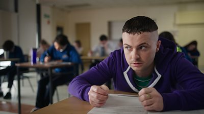 Photo of Conor (Alex Murphy) during filming for The Young Offenders. Sat in a classroom, he leans against his desk and holds a pen above a piece of paper. He looks around the room anxiously. Other students at desks are visible in the background.
