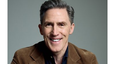 Headshot of Rob Brydon smiling to the camera. He wears a brown jacket and stands in front of a grey background. 